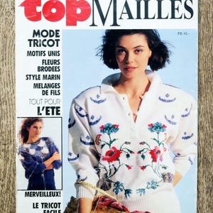 May include: A magazine cover featuring a woman wearing a white knitted sweater with embroidered flowers. The magazine is titled "top MAILLES" and the text "MODE TRICOT" is visible. The cover also includes the text "MERVEILLEUX! LE TRICOT FACILE".