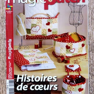Op de afbeelding: Een Franse tijdschriftcover met de titel "magicpatch Hors-Serie" met de woorden "Histoires de coeurs" en de tekst "Panneaux, coussins, objets déco, nécessaire de couture..." De cover toont een verscheidenheid aan patchwork-stoffenartikelen in rood, wit en geel, waaronder een hartvormige tas, een mand en een naaibox.