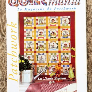 Peut inclure: La couverture du magazine "Quiltmania" présente un motif de patchwork coloré. Le titre du magazine est dans une police multicolore de style patchwork. La couverture comprend également le texte "Le Magazine du Patchwork" et "Les Noeuds Papillons".