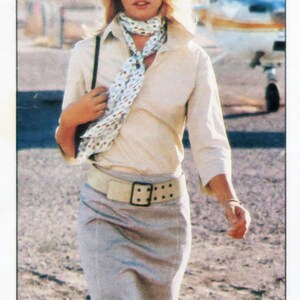 Peut inclure: Une femme portant une chemise blanche avec une écharpe à motifs, une ceinture marron et une jupe grise. Elle porte des lunettes de soleil et se tient devant un petit avion.