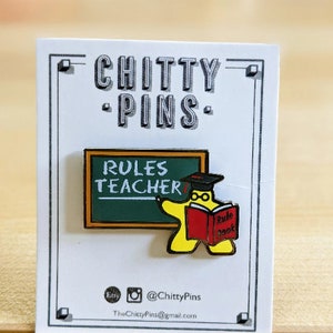 May include: Yellow cartoon teacher holding a red book with the words "Rule Book" on it. The teacher is standing in front of a green chalkboard with the words "Rules Teacher" written on it. The pin is part of the Chitty Pins collection.