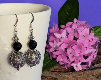 May include: Silver earrings with black beads and ornate silver accents. The earrings are hanging from a white surface.