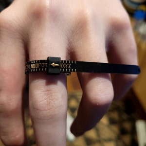 Puede incluir: Un medidor de talla de anillo de plástico negro con letras y marcas doradas. El medidor está marcado con letras de la A a la Z.