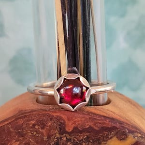 May include: A silver ring featuring a large, round, deep red gemstone. The gemstone is set in a decorative, flower-like bezel. The ring is displayed on a wooden surface, with a glass vase and decorative stems in the background.