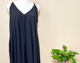 Puede incluir: Vestido negro sin mangas con cuello en V. El vestido se exhibe en un maniquí blanco. El vestido tiene un dobladillo asimétrico y fluido. A la derecha del vestido hay un pequeño taburete redondo de madera con una planta en maceta.