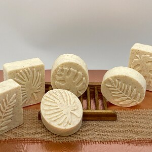 May include: Six handmade soap bars with tropical leaf designs. The soaps are white and have a textured surface. They are arranged on a brown wooden surface with a burlap background.