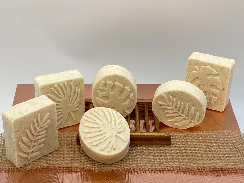 May include: Six handmade soap bars with tropical leaf designs. The soaps are white and have a textured surface. They are arranged on a brown wooden surface with a burlap background.
