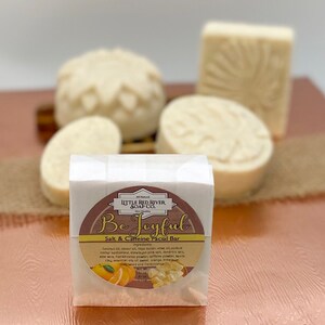 May include: A white bar of soap with a brown label that reads "Be Joyful Salt & Caffeine Facial Bar". The label has a picture of oranges and a small amount of white soap flakes. The soap bar is sitting on a brown surface.