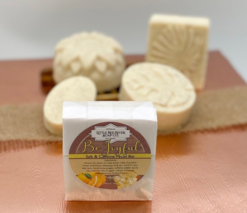 May include: A white bar of soap with a brown label that reads "Be Joyful Salt & Caffeine Facial Bar". The label has a picture of oranges and a small amount of white soap flakes. The soap bar is sitting on a brown surface.