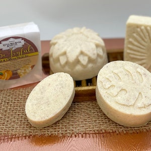 May include: Four white salt and caffeine facial scrub bars with different designs. The bars are on a brown burlap surface. One bar has a leaf design, one has a flower design, one is oval shaped, and one is round with a leaf design. The packaging for the bars is white with the text "Be Joyful" and "Salt & Caffeine Facial Bar".