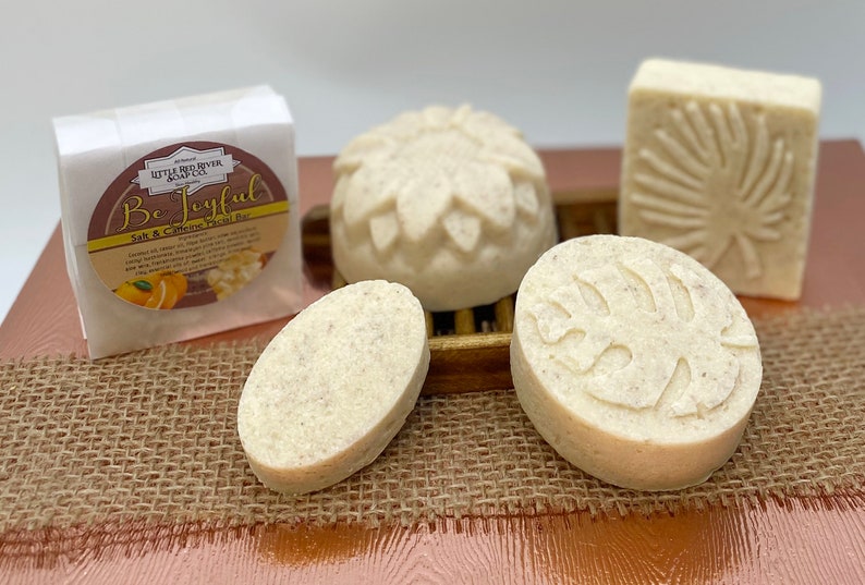 May include: Four white salt and caffeine facial scrub bars with different designs. The bars are on a brown burlap surface. One bar has a leaf design, one has a flower design, one is oval shaped, and one is round with a leaf design. The packaging for the bars is white with the text "Be Joyful" and "Salt & Caffeine Facial Bar".
