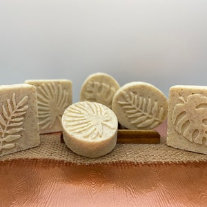 May include: Five handmade soap bars with various tropical leaf designs. The soaps are a light beige color and are arranged on a burlap surface.