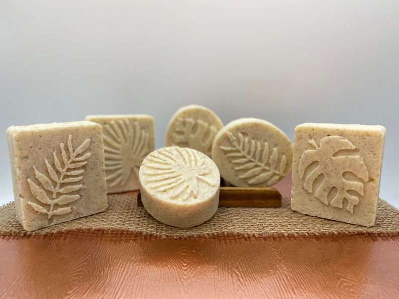May include: Five handmade soap bars with various tropical leaf designs. The soaps are a light beige color and are arranged on a burlap surface.