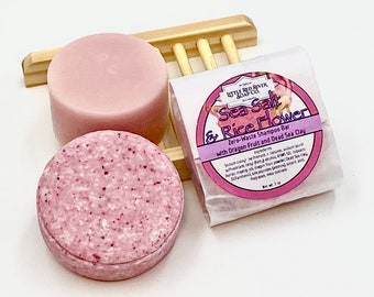 May include: Two pink shampoo bars, one plain and one with a speckled texture, are displayed on a wooden soap dish. The shampoo bar on the right is wrapped in white paper with a pink label that reads "Sea Salt & Rice Flower Zero-Waste Shampoo Bar with Dragon Fruit and Dead Sea Clay".