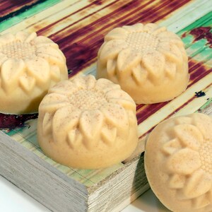 May include: Four handmade soap bars with a sunflower design. The soap bars are a light beige color and are arranged on a wooden surface with a green and brown pattern.