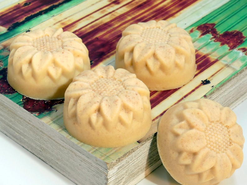 May include: Four handmade soap bars with a sunflower design. The soap bars are a light beige color and are arranged on a wooden surface with a green and brown pattern.