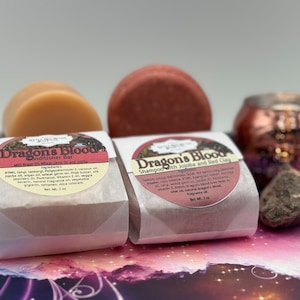 May include: Two white paper-wrapped bars of shampoo and conditioner with the label "Dragon's Blood" and a list of ingredients. The shampoo bar is red and the conditioner bar is beige. The bars are sitting on a purple and pink background.