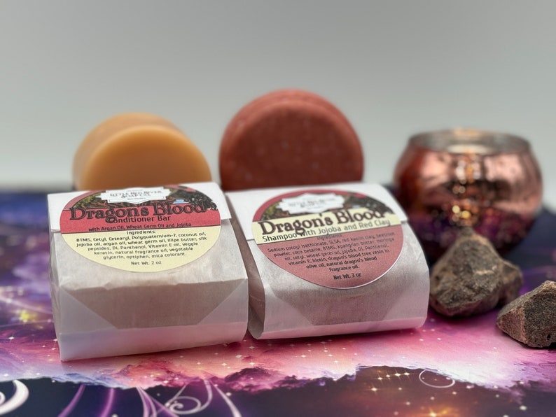 May include: Two white paper-wrapped bars of shampoo and conditioner with the label "Dragon's Blood" and a list of ingredients. The shampoo bar is red and the conditioner bar is beige. The bars are sitting on a purple and pink background.