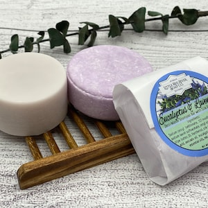 May include: Two round bars of shampoo, one white and one lavender, on a wooden rack. The lavender bar is wrapped in white paper with a blue circle label that reads "Little Red River Soap Co. Eucalyptus & Lavender Zero-Waste Shampoo Bar with Silk Peptides".