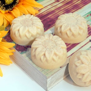 May include: Four round, off-white soap bars with a sunflower design. The soap bars are arranged on a wooden surface with a sunflower in the background.
