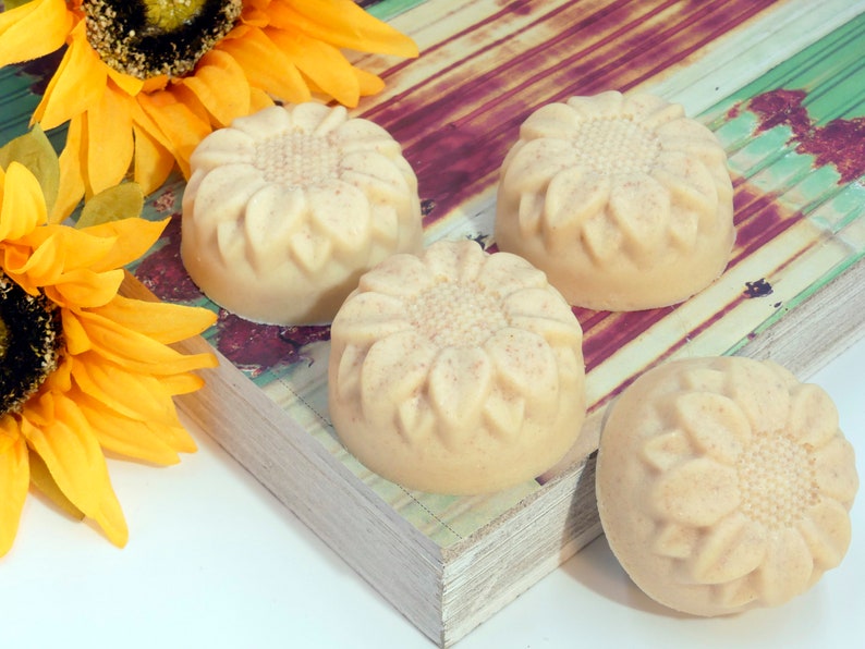 May include: Four round, off-white soap bars with a sunflower design. The soap bars are arranged on a wooden surface with a sunflower in the background.