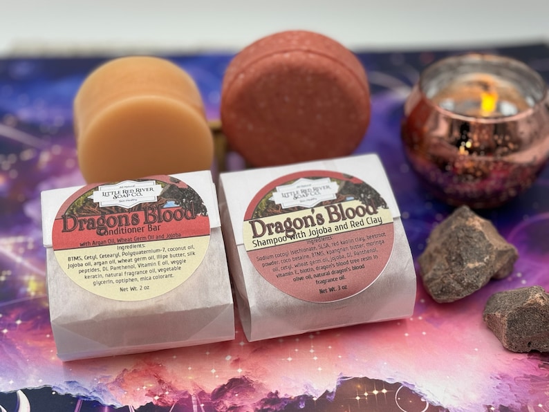 May include: Two white boxes with brown labels containing shampoo and conditioner bars. The labels feature a red circle with a tree and the text "Dragon's Blood". The shampoo bar is labeled "Shampoo with Jojoba and Red Clay" and the conditioner bar is labeled "Conditioner Bar with Argan Oil, Wheat Germ and Jojoba".