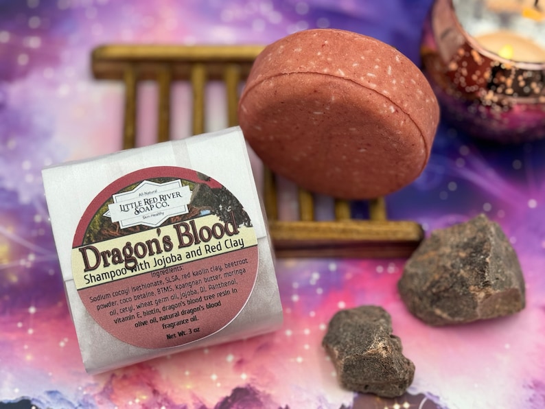 May include: A round, red shampoo bar labeled "Dragon's Blood Shampoo with Jojoba and Red Clay" by Little Red River Soap Co. The bar is sitting on a wooden rack with two small, dark gray rocks in the foreground.