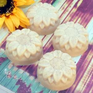May include: Four round, beige-colored soaps with a sunflower design. The soaps are arranged on a striped, wooden surface.