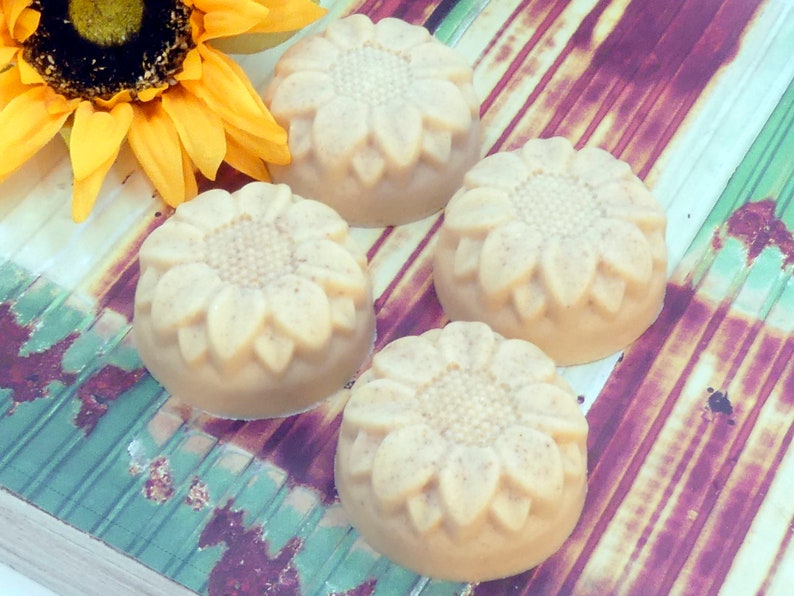 May include: Four round, beige-colored soaps with a sunflower design. The soaps are arranged on a striped, wooden surface.