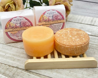 May include: Two bars of soap, one orange and one brown, on a wooden soap dish. The bars are labelled "Amber Rose" and are zero waste shampoo and conditioner bars.
