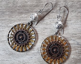 Puede incluir: Un par de pendientes de plata con dijes en forma de engranaje de color dorado. Los dijes tienen un acabado brillante plateado y dorado.