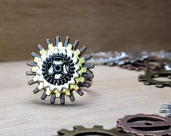 Puede incluir: Un anillo de estilo steampunk plateado y dorado con un diseño de engranaje en capas. El anillo está colocado sobre un fondo de madera con otros engranajes esparcidos a su alrededor.