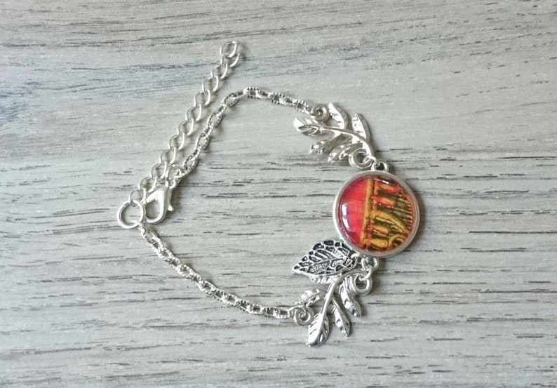 Puede incluir: Pulsera de cadena plateada con un dije redondo de vidrio rojo y amarillo con un dise&ntilde;o de edificio. La pulsera tiene acentos de hojas plateadas.