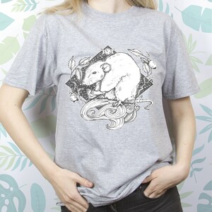 May include: A gray t-shirt with a black and white illustration of a rat with a swirling tail and leaves. The rat is surrounded by a black diamond shape with a moon and stars inside.