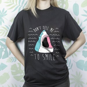 May include: Black t-shirt with a graphic of a shark with its mouth open and the text "Don't tell me to smile".