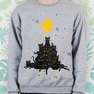May include: A gray sweatshirt with a black cat Christmas tree design. The tree is made up of stacked black cats with yellow eyes and a yellow star on top. The background is a light gray with snowflakes and stars.
