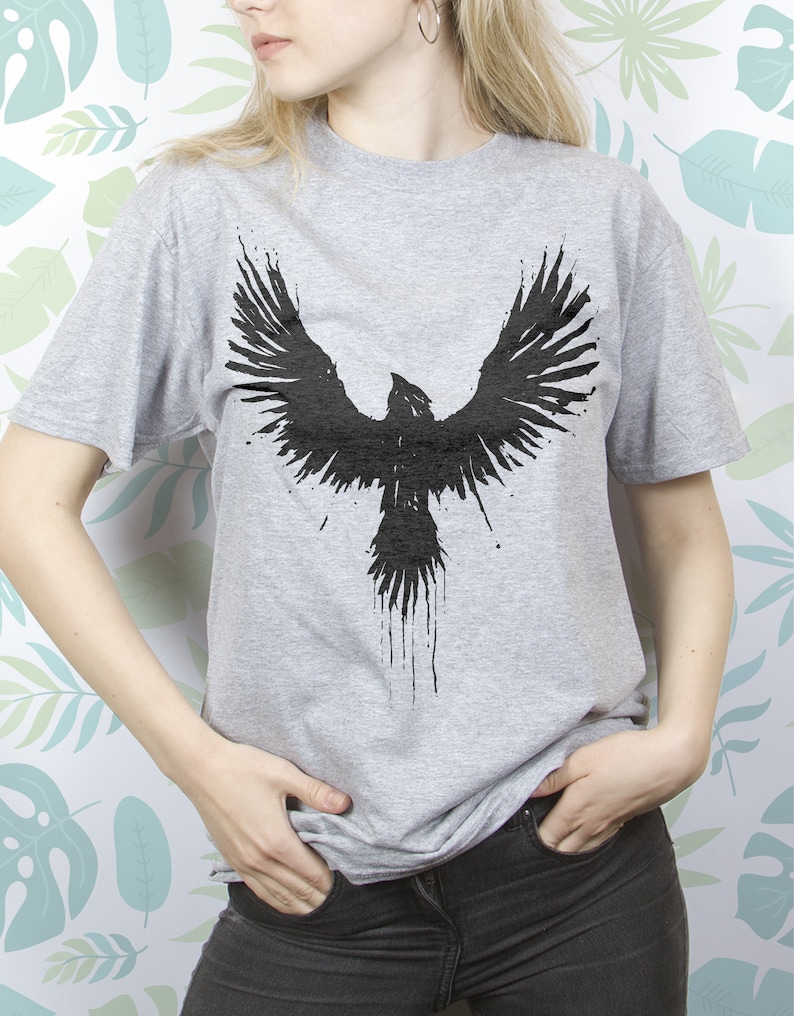 May include: A grey t-shirt featuring a black graphic of a bird with its wings outstretched. The bird is designed to resemble an ink splatter.