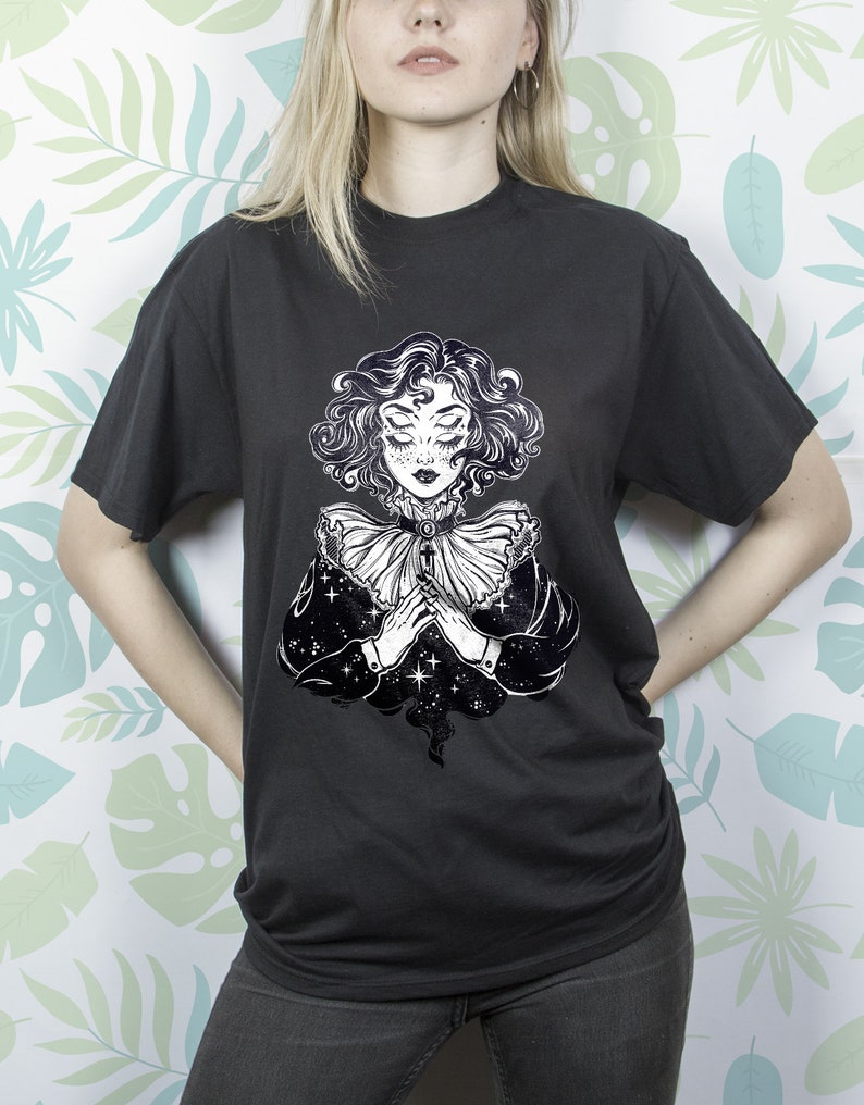 May include: Black t-shirt with a white graphic print of a woman with closed eyes and a large, ruffled collar. The woman's hair is curly and she is wearing a necklace. The background of the graphic is a starry night sky.