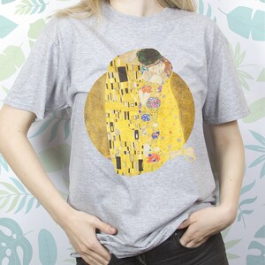 May include: A gray t-shirt with a print of Gustav Klimt's "The Kiss" painting. The painting features two figures embracing in a golden, mosaic-like design.