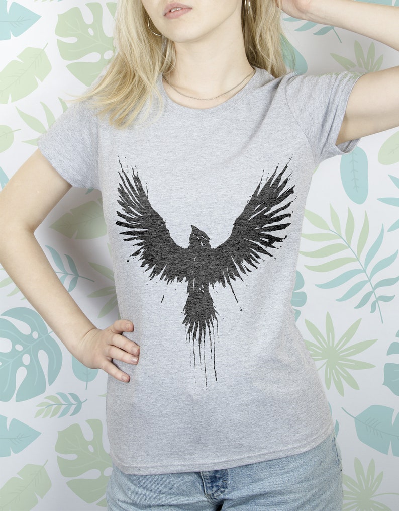 May include: A grey t-shirt featuring a black silhouette of a bird with outstretched wings. The bird is stylised with a dripping paint effect.