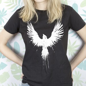 May include: A black t-shirt featuring a white graphic of a bird with its wings outstretched. The bird is depicted in a splatter paint style.