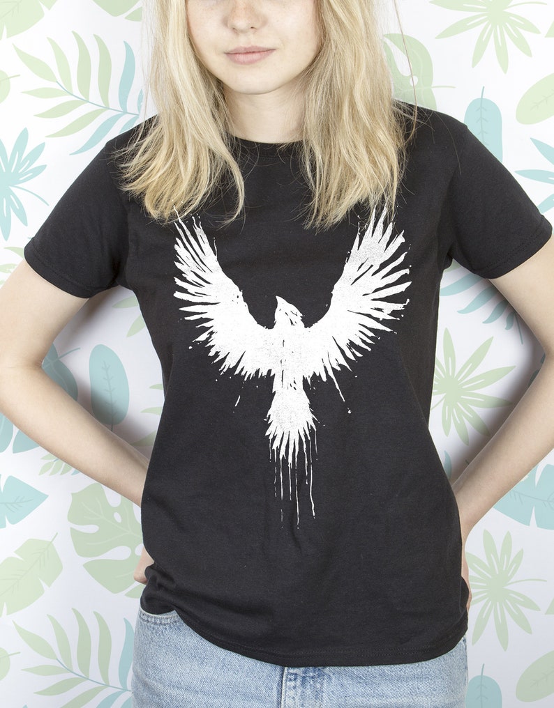 May include: A black t-shirt featuring a white graphic of a bird with its wings outstretched. The bird is depicted in a splatter paint style.