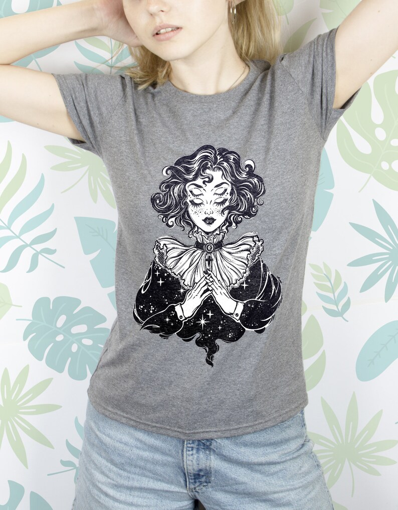May include: Gray t-shirt with a black and white illustration of a woman with multiple eyes and a star-filled sky behind her.