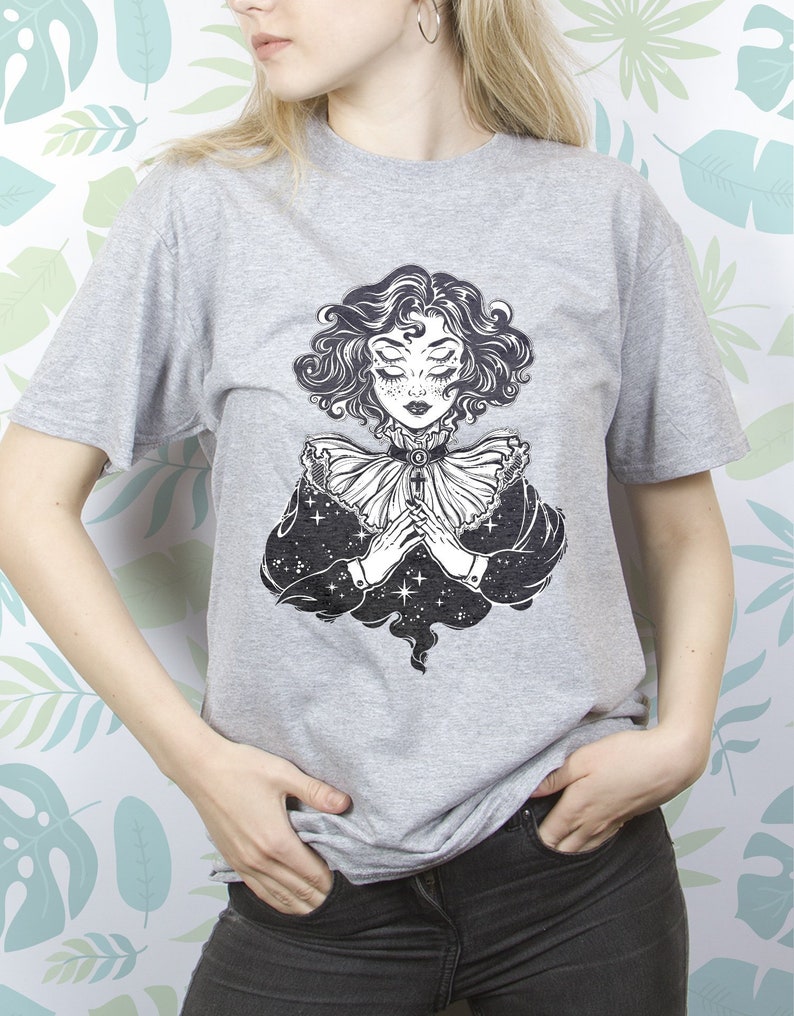May include: A gray t-shirt with a black and white illustration of a woman with her hands clasped together. The woman has a star-shaped pattern on her face and is surrounded by a cloud of stars.