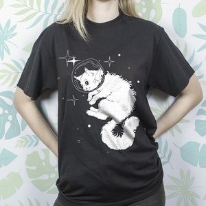 以下が含まれることがあります： 宇宙服を着て宇宙を漂う猫の白いグラフィックがプリントされた黒いTシャツ
