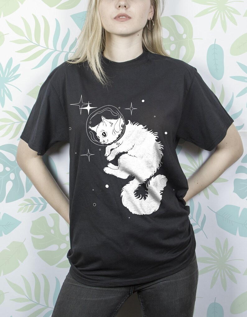 以下が含まれることがあります： 宇宙服を着て宇宙を漂う猫の白いグラフィックがプリントされた黒いTシャツ