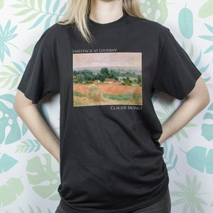 Peut inclure: T-shirt noir avec un imprimé du tableau de Claude Monet, Meules à Giverny. Le tableau représente un champ de meules de foin avec une ferme à l'arrière-plan.