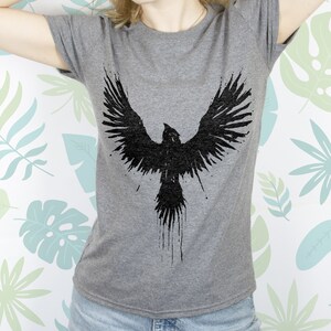 May include: A grey t-shirt featuring a black graphic of a bird with its wings outstretched. The bird is depicted in a splatter style.