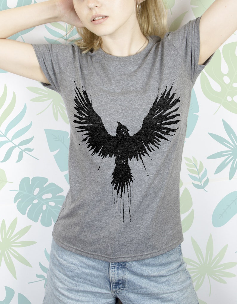May include: A grey t-shirt featuring a black graphic of a bird with its wings outstretched. The bird is depicted in a splatter style.