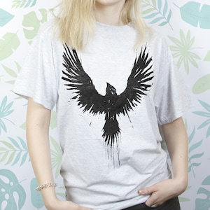 May include: A grey t-shirt featuring a black graphic of a bird with its wings outstretched. The bird is depicted in a stylised, abstract design.